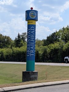 Blue and green Murfreesboro Greenway trailhead marker beside a road.